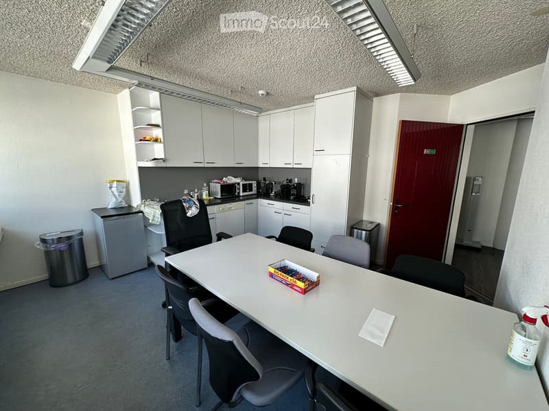 A modern kitchen area with white cabinets, a central table surrounded by chairs, a refrigerator, microwave, and a door leading to another room.