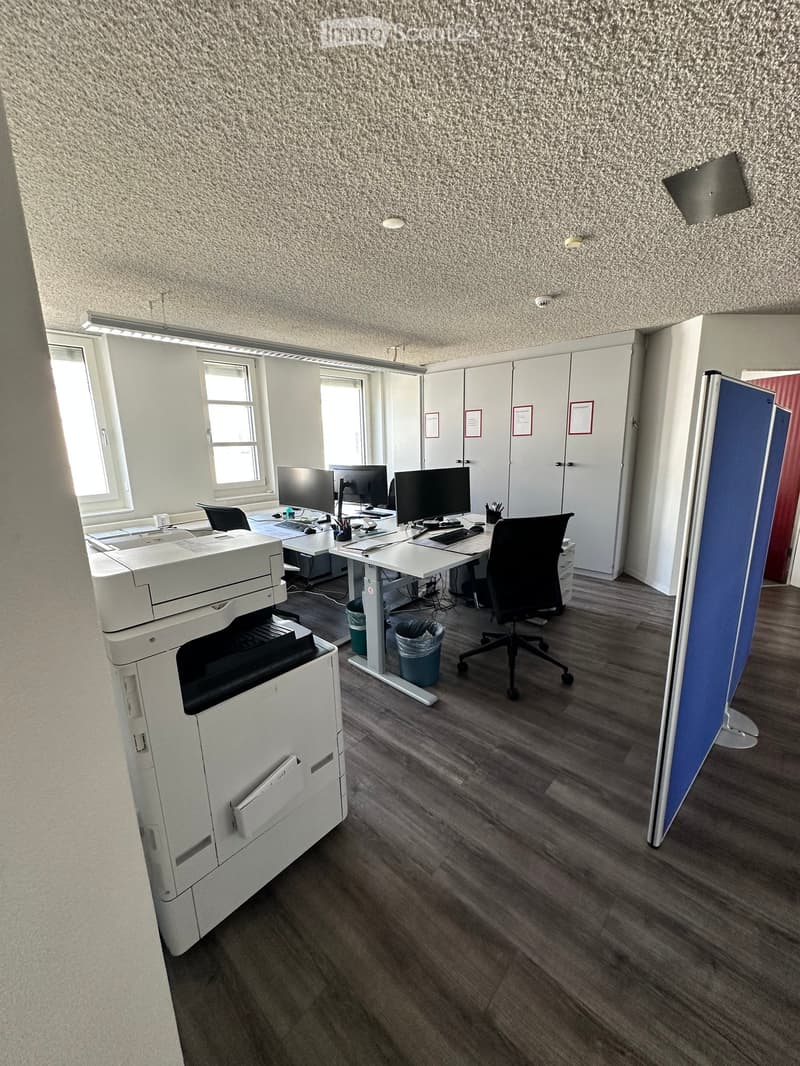 Office space with wooden flooring, white walls, and large windows. Includes desks, chairs, multiple computer screens, a printer, and cabinets.