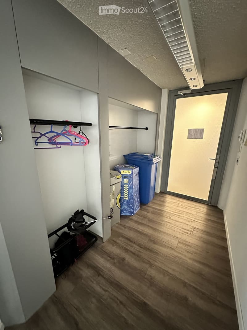 Empty hallway with wooden flooring, white walls, and a door leading to another area. There are storage items like boxes and a stroller in the background.