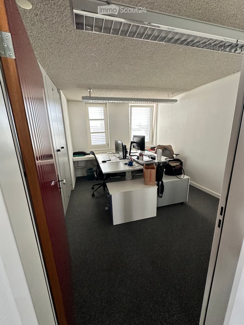 Office space with a desk, chair, computer, and various office supplies. The room has carpeted flooring and white walls.