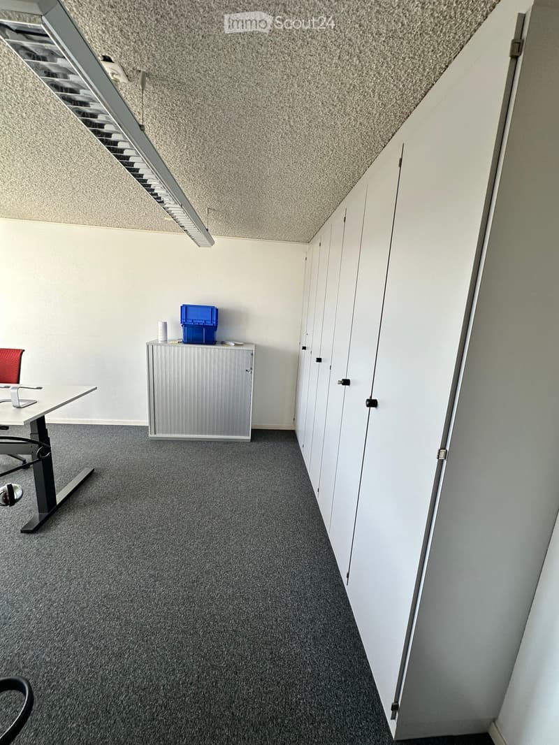 Office space with a grey carpeted floor, white walls, a grey cabinet, a white table with a red chair, and a blue storage box on top of the cabinet.
