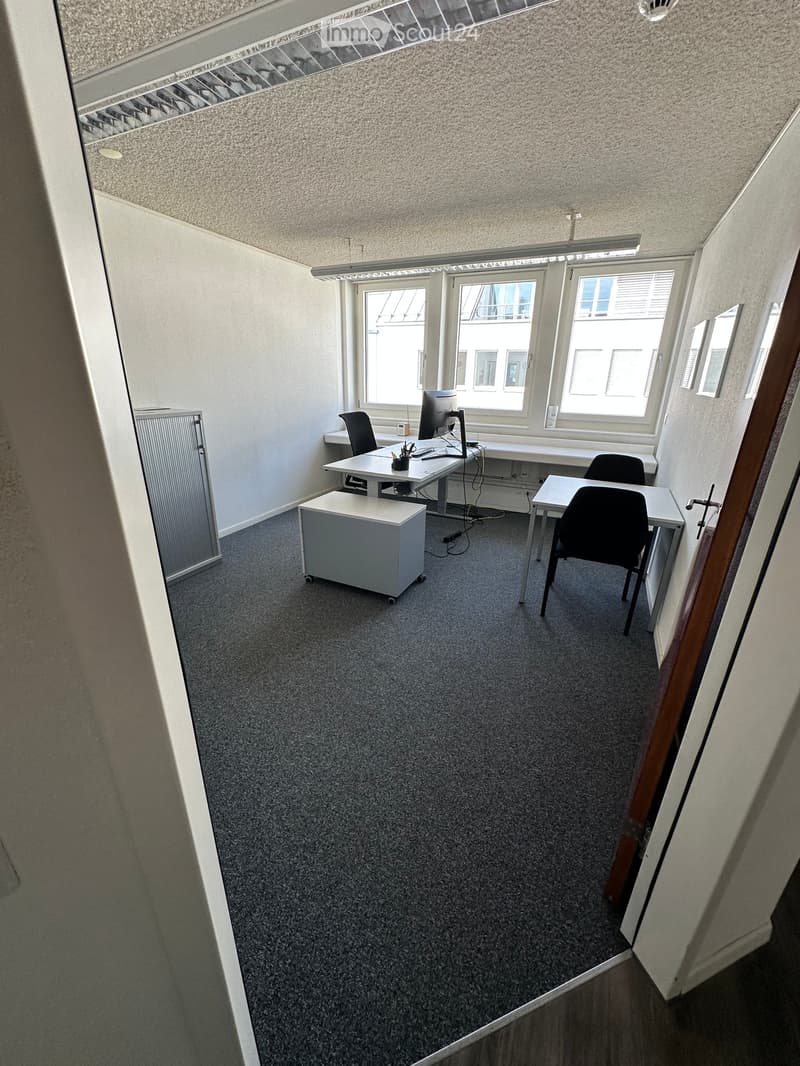 Office space with carpeted floor, white walls, large windows, and modern furniture including desks, chairs, and a computer screen.