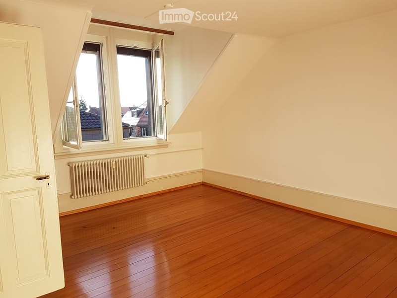 Empty room with wooden flooring, white walls, a radiator, and large windows offering a view of rooftops.