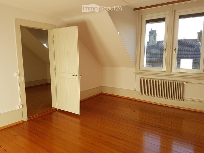 Empty attic room with wooden flooring, white walls, large windows showing a view of rooftops, and a radiator under the windows.