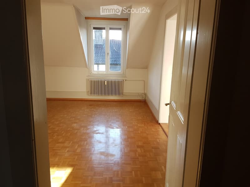 Empty attic room with wooden flooring, white walls, and a window overlooking a roof. A radiator is visible below the window.