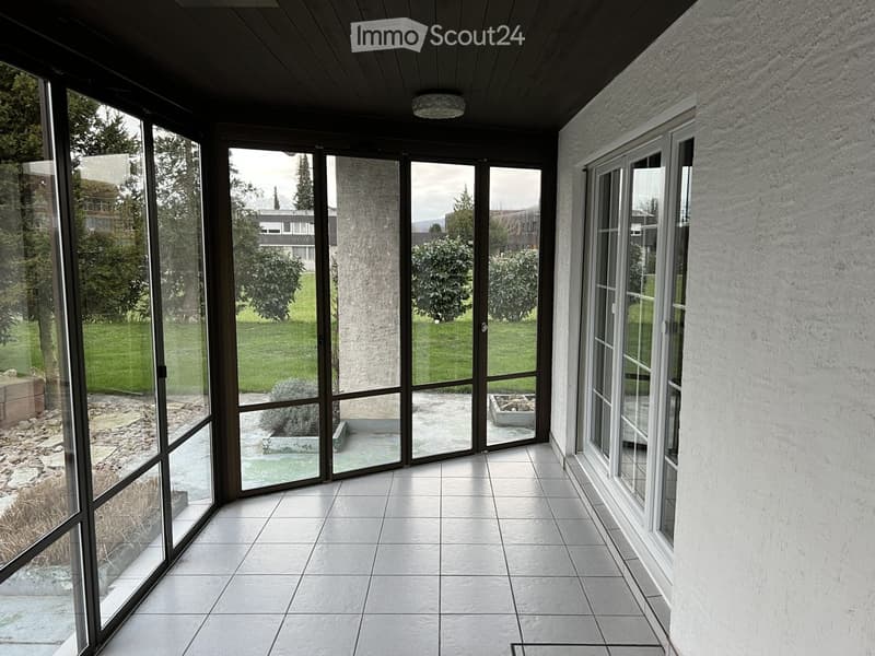 Bright, enclosed balcony with large glass windows and doors, tiled floor, and a view of a landscaped garden.