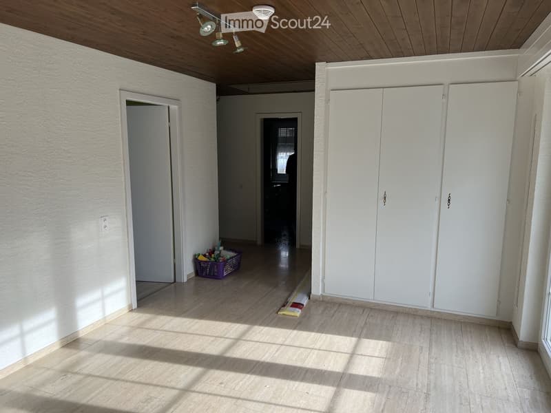 Empty hallway with white walls, wooden ceiling, and tiled floor. Features built-in white cabinets on one side and a small purple basket with cleaning supplies on the floor.
