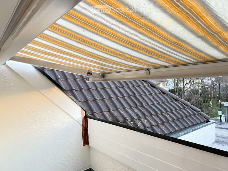 Balcony with a retractable roof, overlooking a tiled roof and surrounding area with trees and a street.