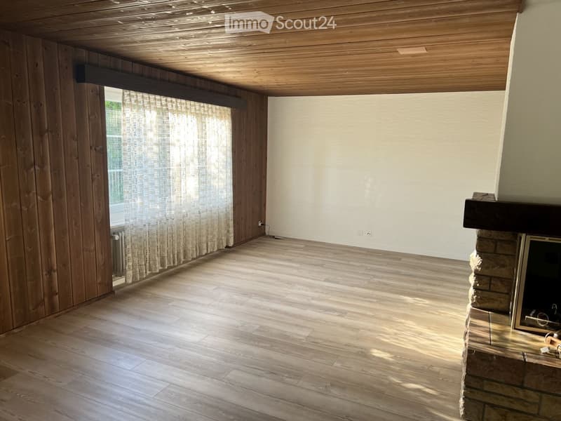 Empty room with wooden walls, wooden ceiling, and wooden floor. Features a fireplace with a stone surround and a window with sheer curtains.