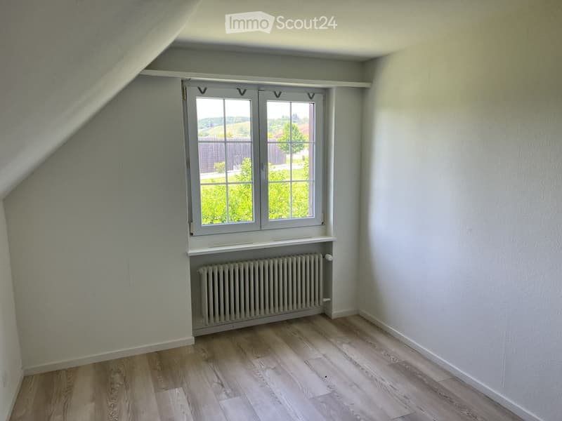 Empty room with light grey walls, wooden flooring, a window with white frame, and a radiator below the window.