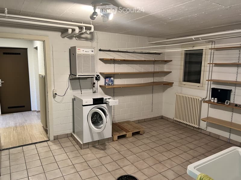A clean, tiled bathroom with a washing machine, radiator, shelves, and an air conditioner. The room features white brick walls, wooden shelves, and a small window.