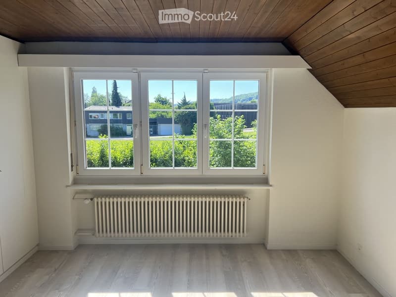 Empty room with wooden flooring, white walls, large window with white frame, radiator below window, and wooden slanted ceiling.