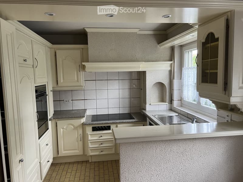 Bright kitchen with cream-colored cabinets, granite countertops, and stainless steel appliances including a sink and oven. The space features a window with a curtain and a decorative backsplash.