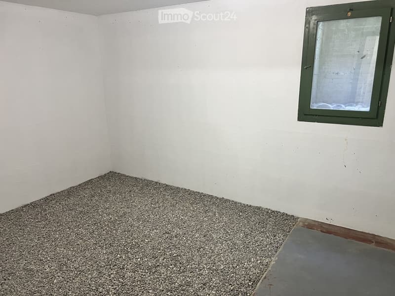 Empty room with white walls, a small window with a green frame, and a gravel floor.