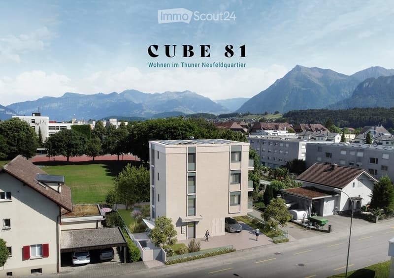A modern multi-story residential building labeled "CUBE 81" in a suburban area with surrounding houses, green spaces, and mountain views.