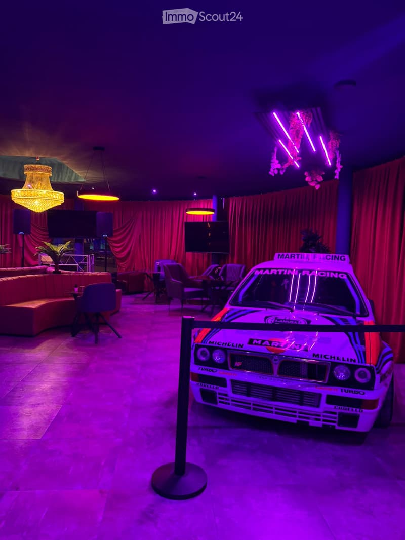 A stylishly furnished room with a vintage racing car prominently displayed, featuring a mix of seating areas, a television, and decorative lighting.