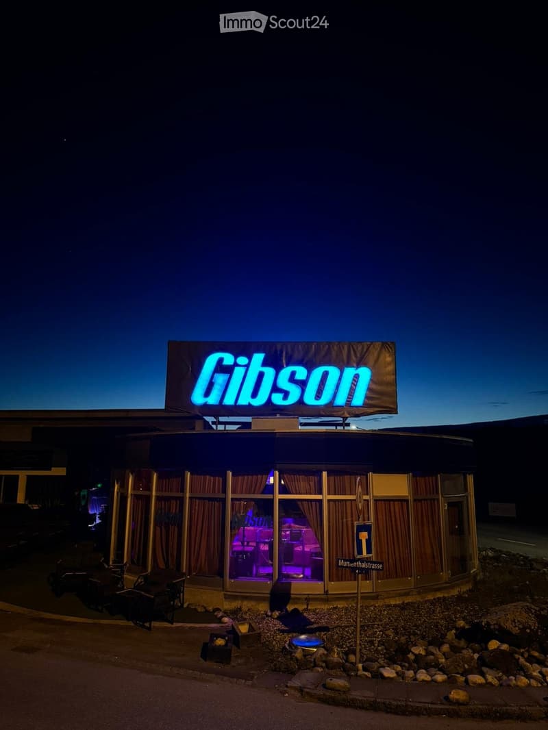 Nighttime exterior view of a building with a neon "Gibson" sign on top, illuminated windows, and a small seating area with chairs and tables outside.
