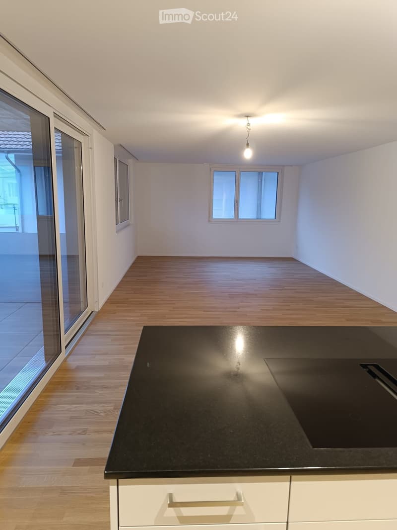 Modern kitchen with a black countertop, white cabinets, and wooden flooring. Large windows and a sliding glass door provide natural light and access to the outdoor area.