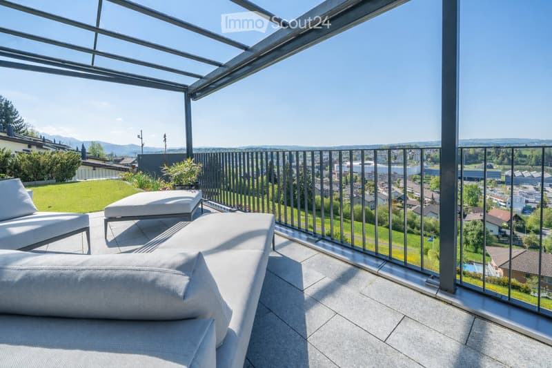 Modern balcony with glass roof and metal railing, featuring white outdoor furniture including a sofa and chairs, overlooking a cityscape with greenery and distant mountains.