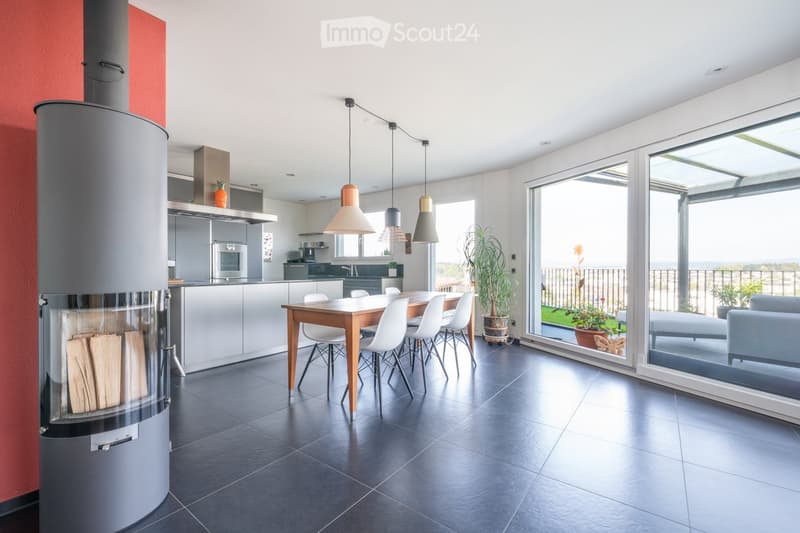 Modern open-plan living and dining area with a central dining table, white chairs, a fireplace, a kitchen area with cabinets and appliances, and large windows overlooking a balcony with outdoor seating.