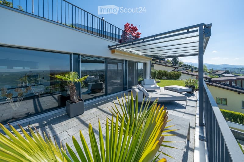 Modern rooftop terrace with a glass railing, outdoor seating area, and potted plants, overlooking a scenic landscape.