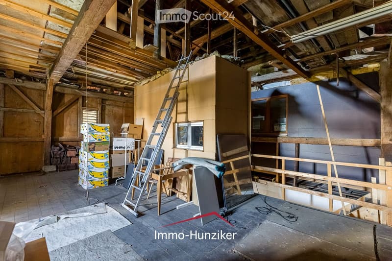 Empty attic space with exposed wooden beams, unfinished walls, and a ladder leading to the upper level. The floor is concrete, and there are some stacked boxes and construction materials visible.