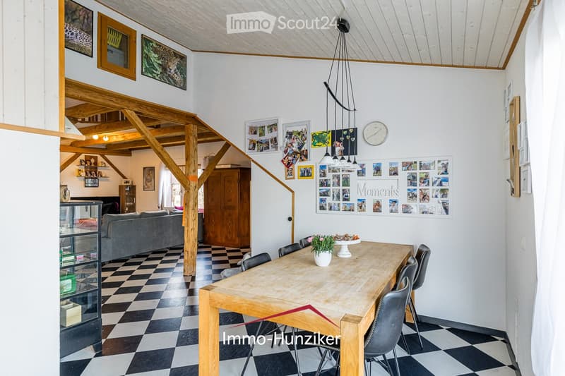 Open-plan living and dining area with exposed wooden beams, checkered black and white flooring, a grey sofa, a wooden dining table with chairs, and a vase with flowers on the table. The walls are decorated with framed photos and artwork.
