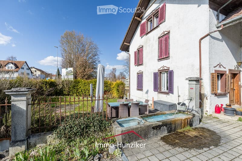 A well-maintained garden area with a small fountain, a table and chairs, and a stone pathway leading to a two-story house with red shutters and a wooden door.