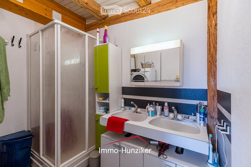 Modern bathroom with double sink, mirrored cabinet, shower, and various toiletries. Features exposed wooden beams on the ceiling and a mix of white and blue wall tiles.