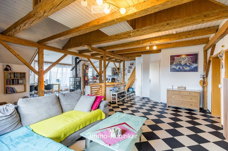 Open-concept living and dining area with exposed wooden beams, checkered flooring, a grey sofa with colorful cushions, a glass coffee table, and a bookshelf. The space is connected to a modern kitchen area with a counter and bar stools.