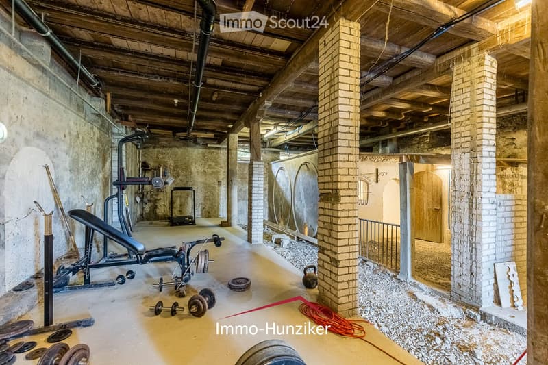 A spacious, unfinished basement gym with exposed brick pillars, wooden beams, and concrete floors. Contains a bench press, dumbbells, weight plates, a cable machine, and a weight rack.