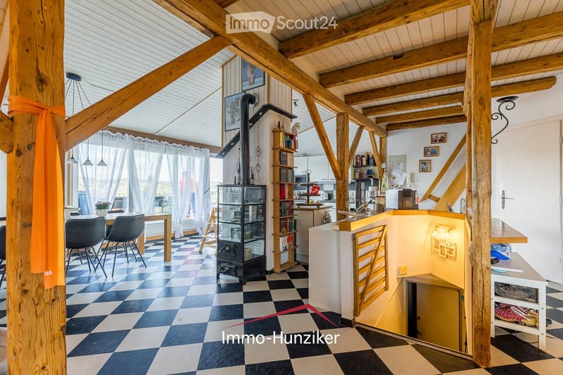 Open-plan living and dining area with exposed wooden beams, checkered flooring, and a modern kitchen area. Features include a dining table with chairs, a kitchen counter, a bookshelf, and a refrigerator.