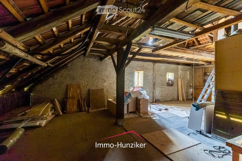 An attic space with exposed wooden beams and a slanted ceiling, featuring a concrete wall, a small window, and scattered construction materials and tools.