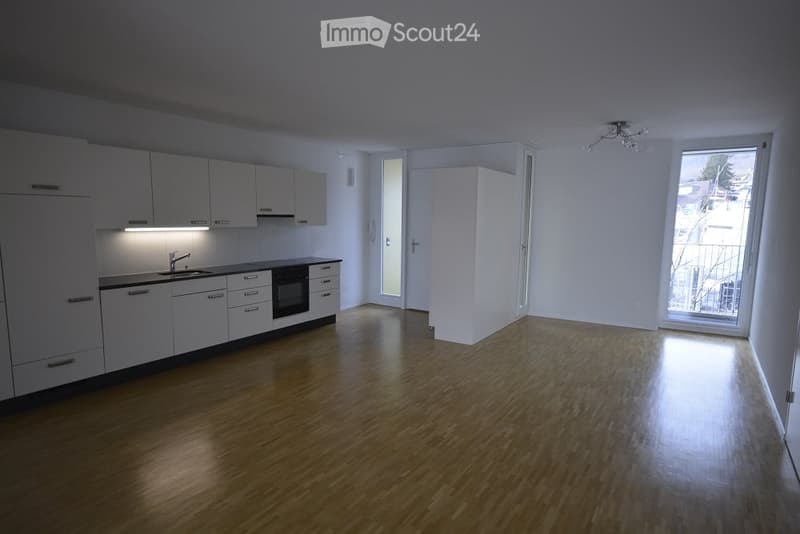 Empty room with wooden flooring, white walls, and a small kitchen area with white cabinets and a sink.