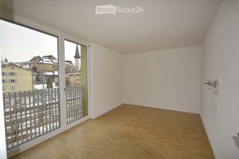 Empty room with wooden flooring, large window offering a view of a town with a church tower, and a metal security gate visible outside.