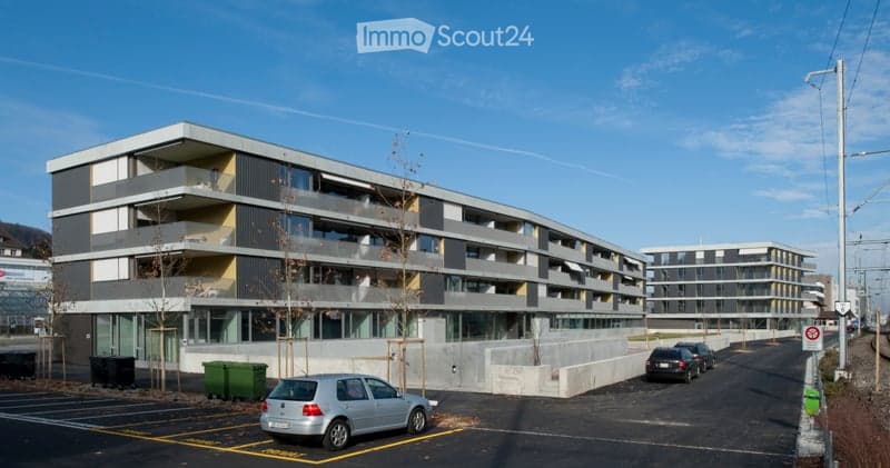Modern multi-story residential building with balconies, large windows, and a parking area.