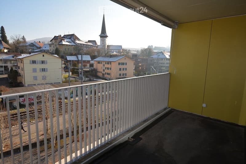 Balcony with white railing overlooking a town with houses and a church tower in the background.