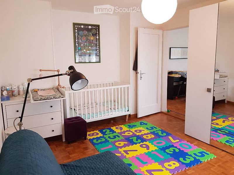 Brightly lit bedroom with a crib, colorful play mat, and a desk area visible through an open door.