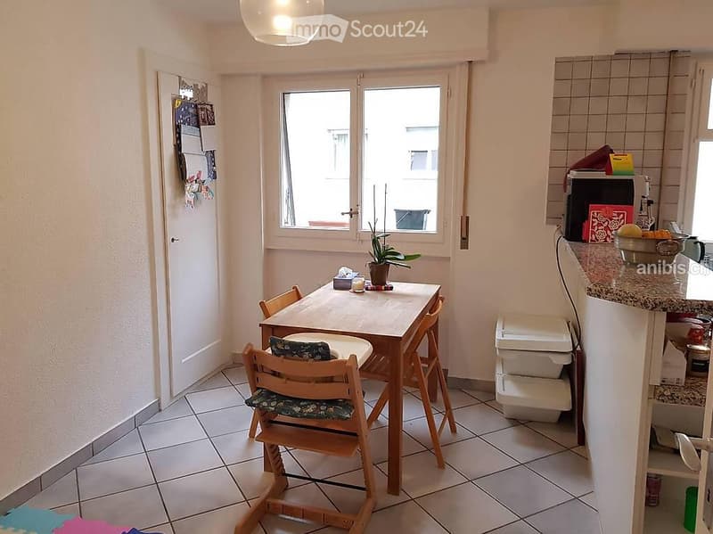 A small, well-lit dining area with a wooden dining table and chairs, a potted plant on the table, and a microwave on the counter.
