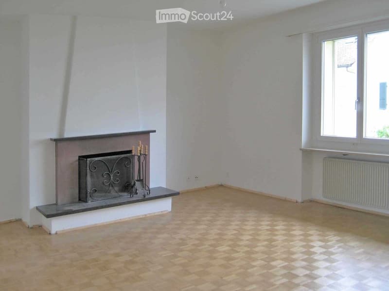 Empty room with wooden flooring, white walls, a fireplace with a decorative screen, and a window with a radiator below it.