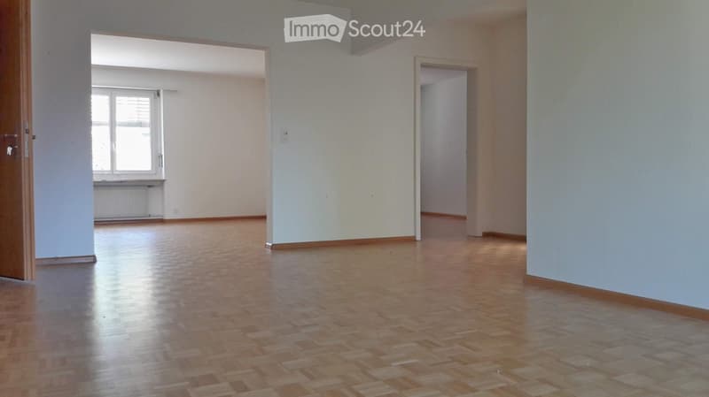 Empty room with wooden flooring, white walls, and a large window with blinds. The space is open and well-lit, with no furniture or decorations present.