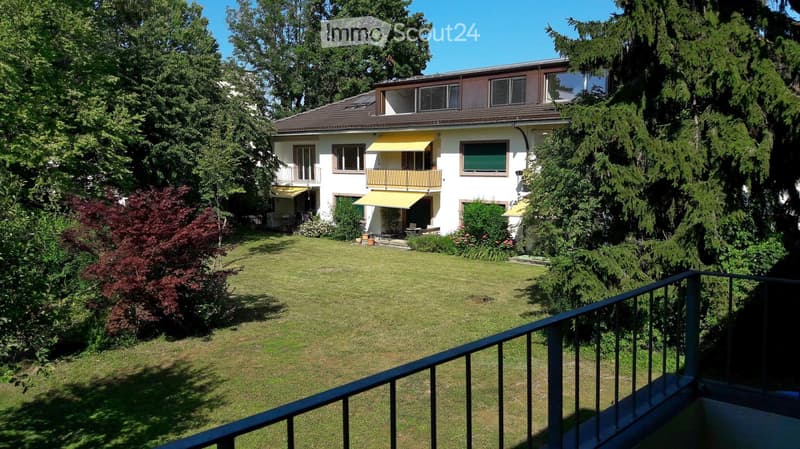 A well-maintained garden with a lawn, surrounded by trees and shrubs, with a white house featuring balconies and yellow awnings in the background.
