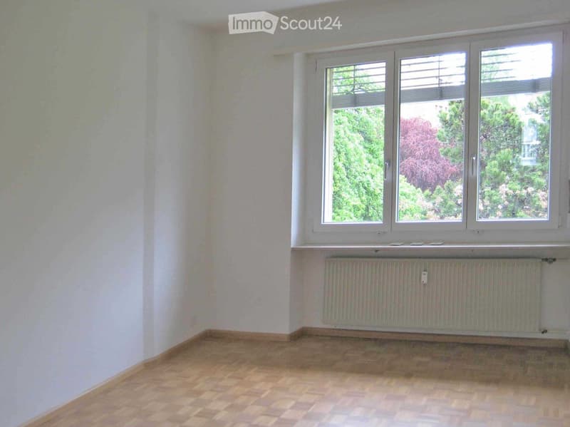 Empty room with wooden flooring, white walls, and a large window with blinds. A radiator is visible below the window.