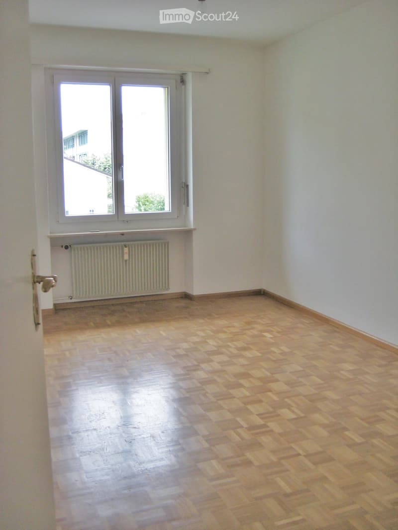 Empty room with wooden flooring, white walls, a radiator, and a large window with double doors leading outside.