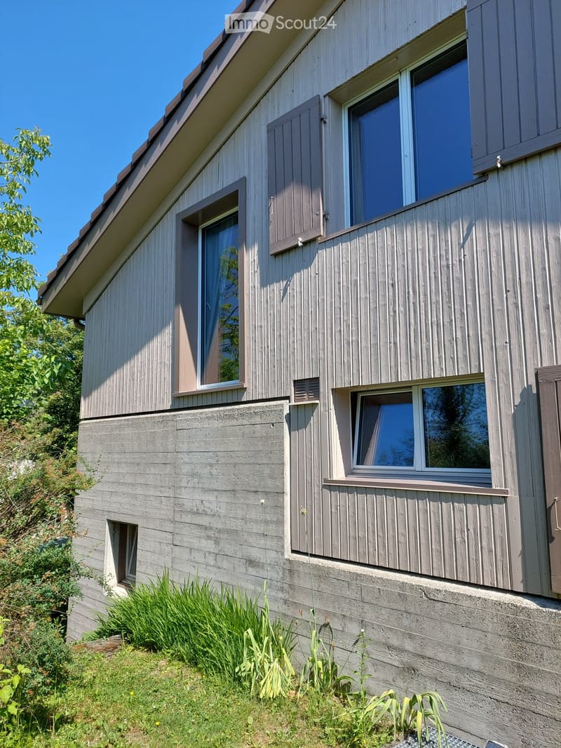 Modern two-story house with a combination of wood siding and concrete walls, featuring multiple windows with shutters and a small raised ground floor entrance.