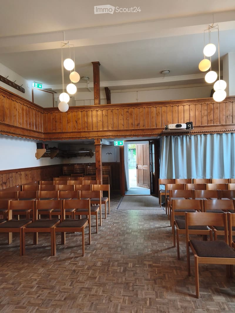 Wooden chairs arranged in rows, wooden panel walls, hanging spherical lights, projector and speakers on a wooden shelf, and a doorway leading outside.