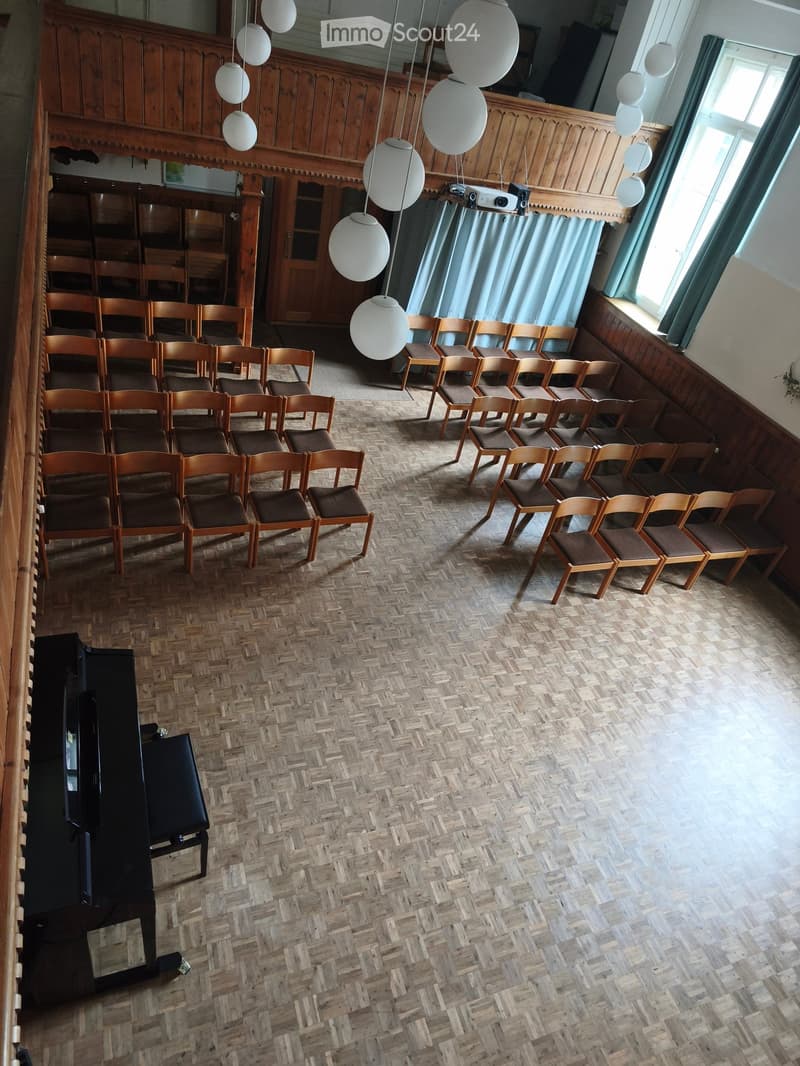 Wooden chairs arranged in rows, a grand piano, wooden walls, and large windows with curtains.