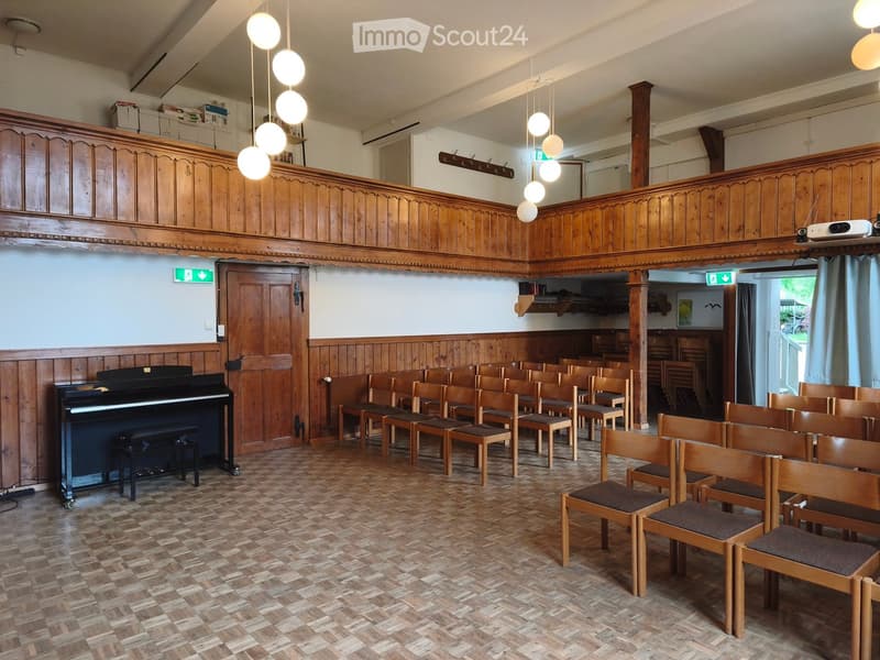 Wooden-paneled room with rows of chairs, a piano, and a projector on a shelf. Wooden floor and decorative wooden trim on walls.