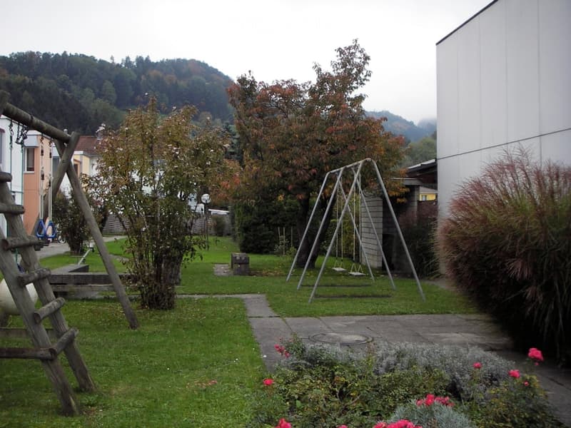 A well-maintained garden with a swing set, climbing frame, and various plants and trees, surrounded by a paved pathway and a modern building.