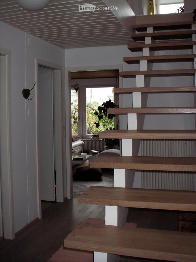Modern hallway with wooden staircase leading to another room, visible furniture in the adjacent space includes a sofa and coffee table.